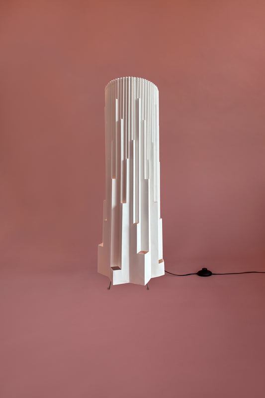 Lámpara de piso de Colombia. Plié Lights, referencia  Torre de la colección monumentos. Fotografía de producto confondo de color rosa. Caperuza escultórica estílo arquitectónico. El papel tiene cortes lo que permite genera movimiento entre luz directa e indirecta. Hecha en papel a mano. La base metálica es color negro y cable negro.