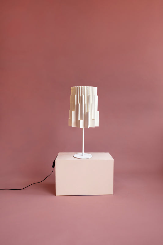 Plié Lights, lámpara de mesa referencia Pilar, de la colección Monumentos, hecha a mano, caperuza hecha en papel plegado a mano. Soporte metálica y cable blanco Acabado negro. Producto hecho en Colombia. Diseño Colombiano. Figura geométrica muy interesante. Ideal para iluminar y darle calidez a los espacios. efecto de luz y sombras generado por cortes en el papel que dejan pasar luz directa.