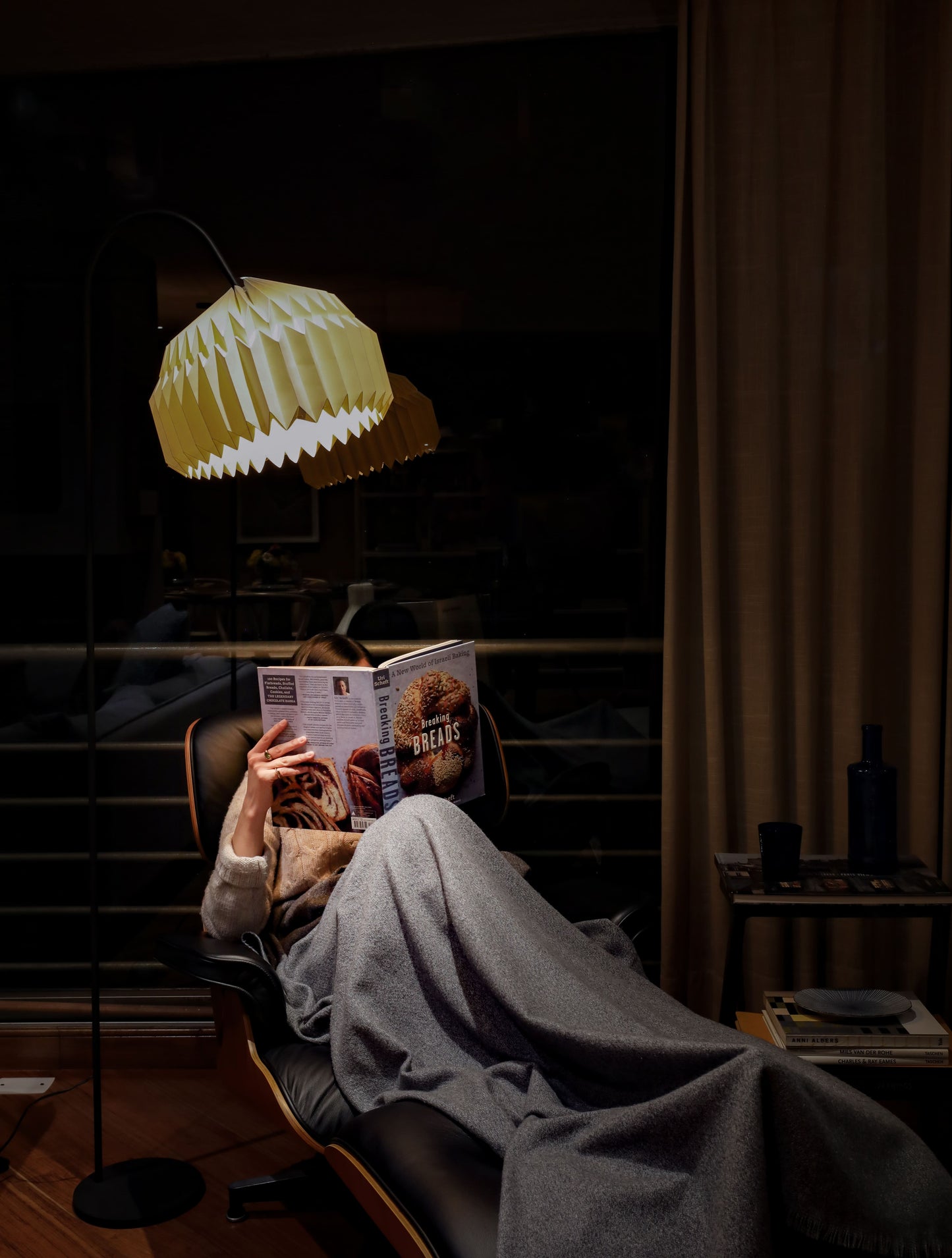 Plié Lights, lámpara de piso en forma de arco hecha en colombia. La base metálica tiene un tamaño perfecto para lámpara de lectura. La caperuza es referencia Alfa, tiene un diseño que genera luz directa hacia abajo. El diseño tiene pliegues en papel geométricos. Produce una iluminación cálida. Fotografía de producto en sitio, la lámpara está en un estudio, es de noche y la lámpara esta prendida, iluminando a una persona mientras que está sentada en una silla lounge, con una cobija y un libro