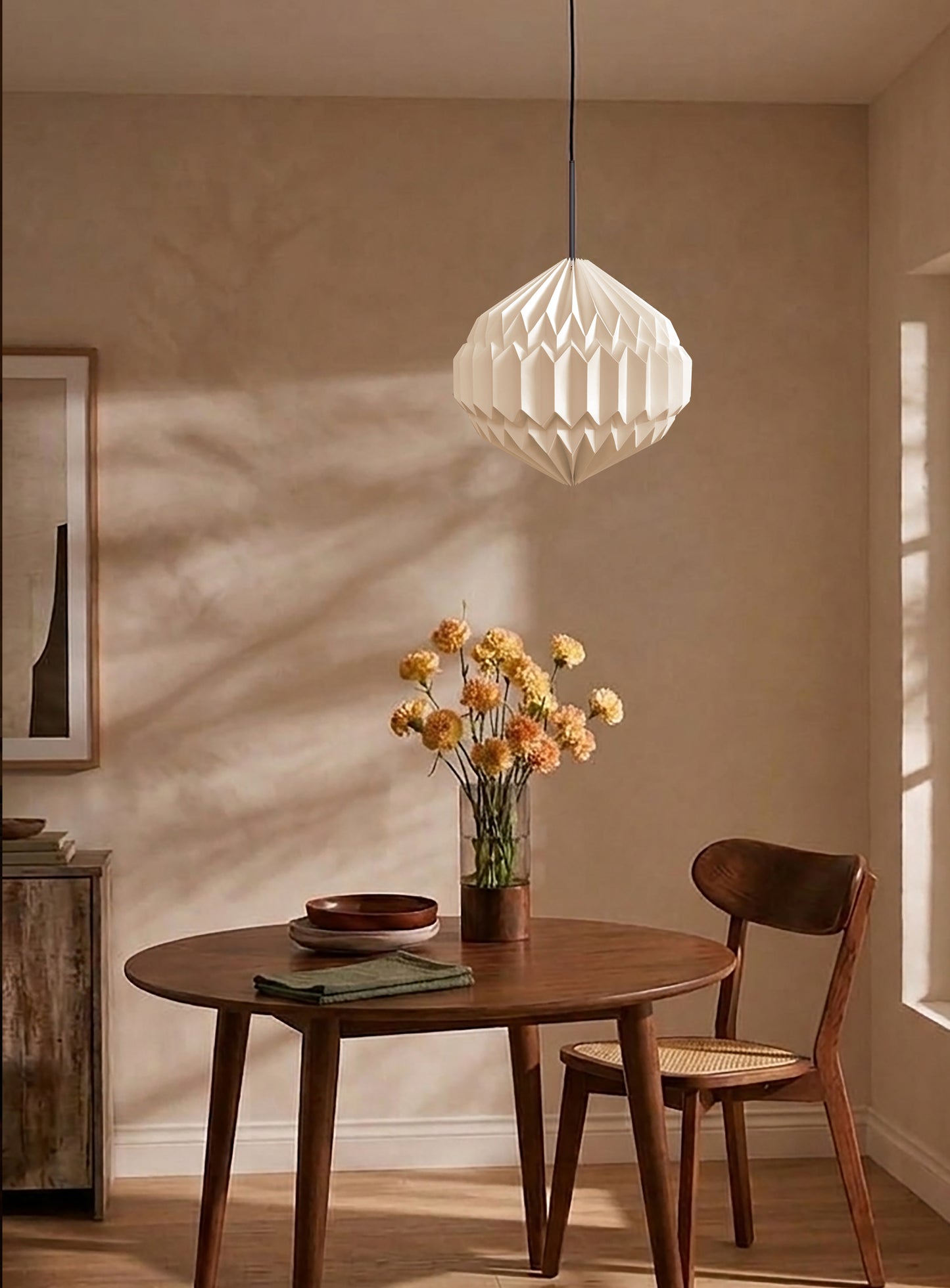 Plié Lights, lámpara colgante referencia luna, hecha a mano, caperuza hecha en papel plegado a mano. Soporte metálica y cable negro Acabado negro. Producto hecho en Colombia. Diseño Colombiano. Figura geométrica muy interesante. Ideal para iluminar y darle calidez a los espacios. Fotografía de producto colgada encima de una mesa de comedor. Sobre la mesa hay un florero, unos platos y la escena es de día y la lámpara está apagada.