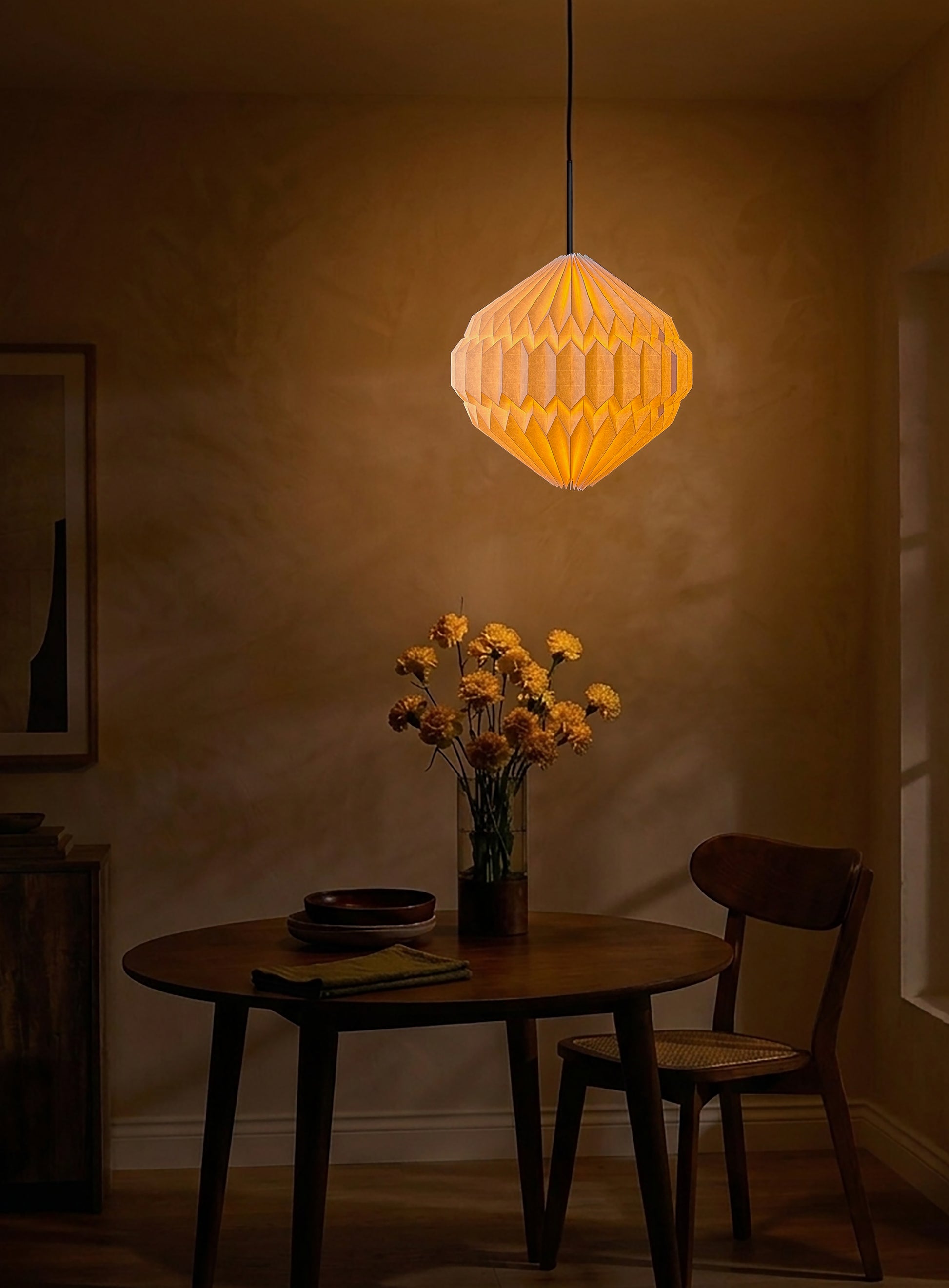 Plié Lights, lámpara colgante referencia luna, hecha a mano, caperuza hecha en papel plegado a mano. Soporte metálica y cable negro Acabado negro. Producto hecho en Colombia. Diseño Colombiano. Figura geométrica muy interesante. Ideal para iluminar y darle calidez a los espacios. Fotografía de producto colgada encima de una mesa de comedor. Sobre la mesa hay un florero, unos platos y la escena es nocturna y el ambiente es cálido. 