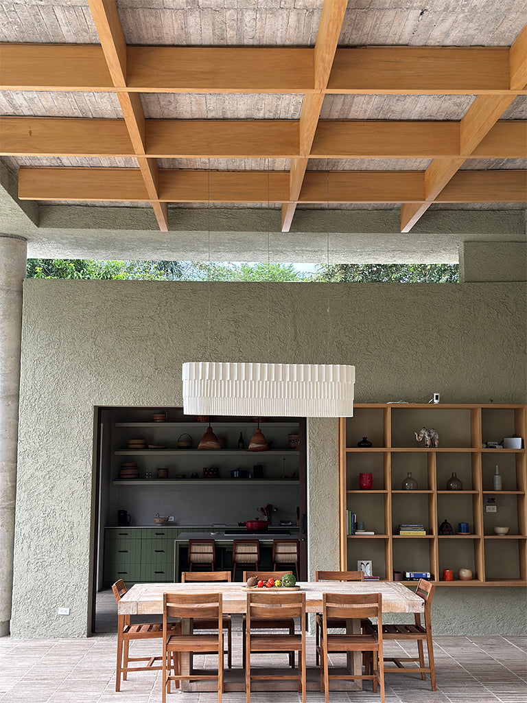 Lámpara colgante de Colombia. Plié Lights, referencia Capitolio de la colección monumentos. Fotografía en sitio, colgada en un comedor, se ve la mesa de comedor con ocho sillas en un espacio abierto, al fondo se ve un jardín, el techo es alto. La lámpara de Plié está apagada y es tomada durante el día. Caperuza escultórica estilo arquitectónico. El papel tiene cortes lo que permite genera movimiento entre luz directa e indirecta. Hecha en papel a mano.