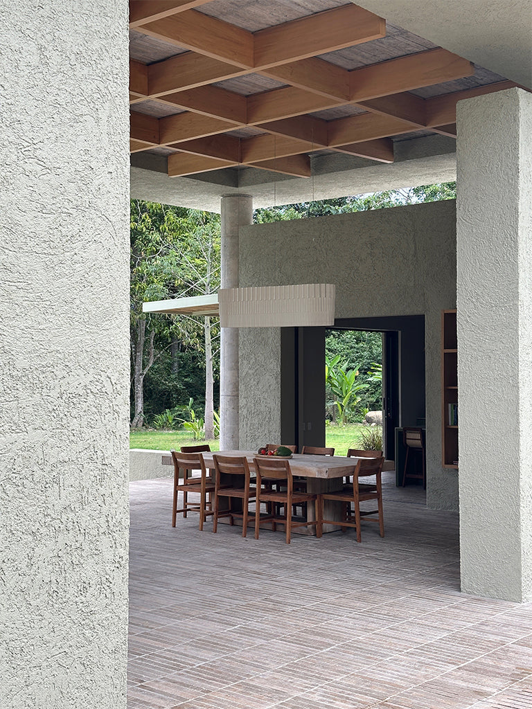 Lámpara colgante de Colombia. Plié Lights, referencia Capitolio de la colección monumentos. Fotografía en sitio, colgada en un comedor, se ve la mesa de comedor con ocho sillas en un espacio abierto, al fondo se ve un jardín, el techo es alto. La lámpara de Plié está apagada y es tomada durante el día. Caperuza escultórica estilo arquitectónico. El papel tiene cortes lo que permite genera movimiento entre luz directa e indirecta. Hecha en papel a mano.