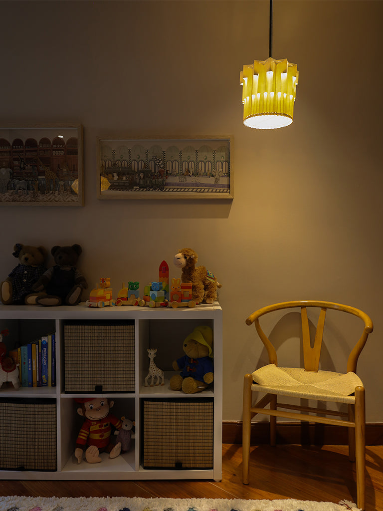 Lámpara colgante de Colombia. Plié Lights, referencia  Fortaleza de la colección Monumentos. Fotografía tomada en sitio, lámpara colgando en un cuarto de niños, se ve una biblioteca baja con juguetes, una silla esquinera y unos cuadros con ilustraciones infantiles. Diseño geométrico inspirado en la arquitectura. Caperuza de papel doblada con crtes que generan una iluminación arquitectónica nocturna. Hecha en papel a mano. La base metálica es color negro y cable negro.