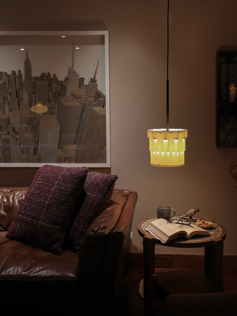 Lámpara colgante de Colombia. Plié Lights, referencia  Fortaleza de la colección Monumentos. Fotografía tomada en sitio, lámpara colgando encima de una mesa auxiliar que está al lado de una sofá. Encima de la mesa hay una libro, una merienda ó snack y un café. Diseño geométrico inspirado en la arquitectura. Caperuza de papel doblada con crtes que generan una iluminación arquitectónica nocturna. Hecha en papel a mano. La base metálica es color negro y cable negro.