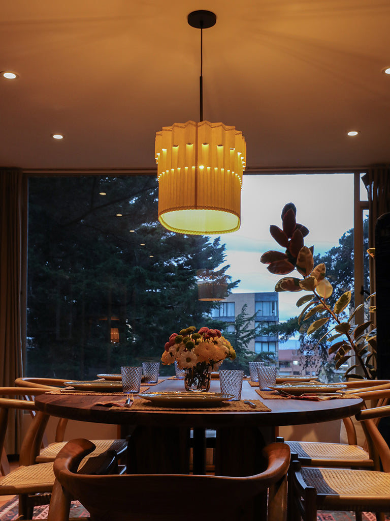 Lámpara colgante de Colombia. Plié Lights, referencia Museo de la colección monumentos. Fotografía en sitio, colgada en un comedor, se ve la mesa de comedor con 5 sillas y la ventana con un pino y un edificio en el fondo. La lámpara de Plié está prendida y es tomada durante el día. Caperuza escultórica estilo arquitectónico. El papel tiene cortes lo que permite genera movimiento entre luz directa e indirecta. Hecha en papel a mano. La base metálica es color negro y cable negro.