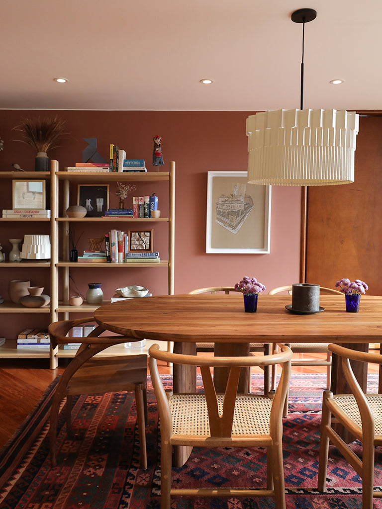 Lámpara colgante de Colombia. Plié Lights, referencia Museo de la colección monumentos. Fotografía en sitio, colgada en un comedor, se ve la mesa de comedor con 5 sillas y una biblioteca con objetos y un cuadro en el fondo. La lámpara de Plié está apagada y es tomada durante el día. Caperuza escultórica estilo arquitectónico. El papel tiene cortes lo que permite genera movimiento entre luz directa e indirecta. Hecha en papel a mano. La base metálica es color negro y cable negro.