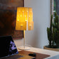 Fotografía de la lámpara Plié Lights, lámpara de mesa referencia Pilar, de la colección Monumentos. Producto hecho en Colombia. Diseño Colombiano. Figura geométrica muy interesante. Ideal para iluminar y darle calidez a los espacios. efecto de luz y sombras generado por cortes en el papel que dejan pasar luz directa. La lámpara está puesta en un escritorio, está prendida, hay un computador en primer plano, en el fondo hay un cuadro y un cactus.
