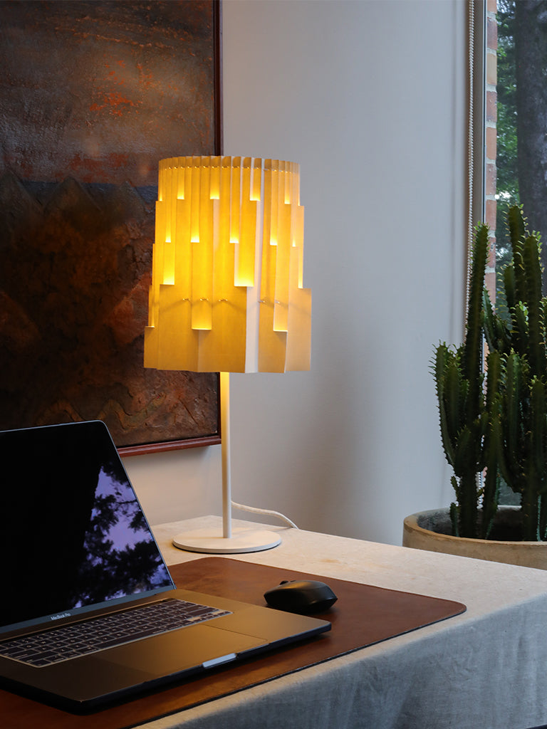 Fotografía de la lámpara Plié Lights, lámpara de mesa referencia Pilar, de la colección Monumentos. Producto hecho en Colombia. Diseño Colombiano. Figura geométrica muy interesante. Ideal para iluminar y darle calidez a los espacios. efecto de luz y sombras generado por cortes en el papel que dejan pasar luz directa. La lámpara está puesta en un escritorio, está prendida, hay un computador en primer plano, en el fondo hay un cuadro y un cactus.