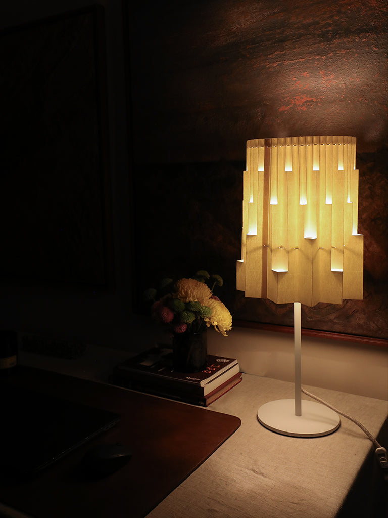 Fotografía de la lámpara Plié Lights, lámpara de mesa referencia Pilar, de la colección Monumentos. Producto hecho en Colombia. Diseño Colombiano. Figura geométrica muy interesante. Ideal para iluminar y darle calidez a los espacios. efecto de luz y sombras generado por cortes en el papel que dejan pasar luz directa. La lámpara está puesta en un escritorio, se ve cómo muy sutilmente se ilumina un florero a mano izquierda.