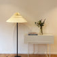 Lámpara de piso marca Plié referencia Andrómeda puesta en una espacio al lado de una mesa.  Fabricada con base metálica y caperiza hecha en papel. Diseño colombiano hecho a mano.