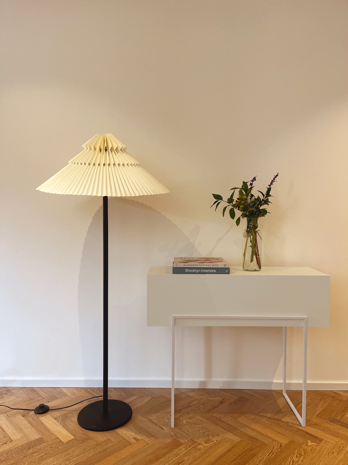 Lámpara de piso marca Plié referencia Andrómeda puesta en una espacio al lado de una mesa.  Fabricada con base metálica y caperiza hecha en papel. Diseño colombiano hecho a mano.