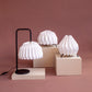 Tres lámparas de mesa marca Plié, referencias Lyra, Fenix y Gemini. Fabricadas con base metálica y caperuzas hechas en papel plegado. Perfectas para iluminar espacios pequeños y dar un brillo a los espacios. Dos lámparas son pequeñas y la otra es tamaño mediano. Diseño colombiano. Hecha a mano en Colombia.
