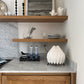 Lámpara de mesa marca Plié, referencia Gemini. Fabricada con base metálica y caperuza hecha en papel plegado. Perfecta para iluminar espacios pequeños y dar un brillo a los espacios. La lámpara esta puesta en el mesón de la cocina para mostrar el tamaño de la lámpara. Diseño colombiano. Hecha a mano en Colombia