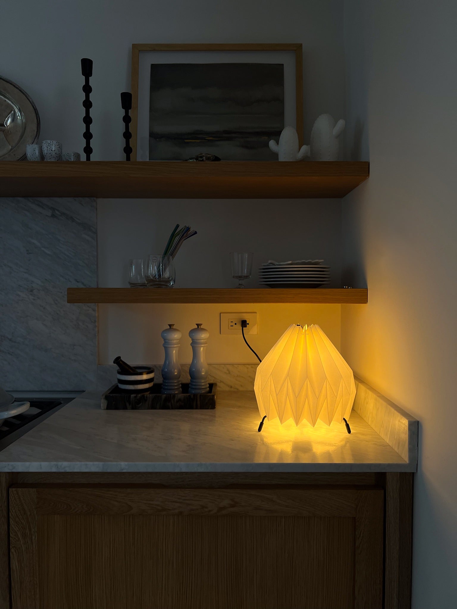 Lámpara de mesa marca Plié, referencia Gemini. Fabricada con base metálica y caperuza hecha en papel plegado. Perfecta para iluminar espacios pequeños y dar un brillo a los espacios. La lámpara esta puesta en el mesón de la cocina para mostrar el tamaño de la lámpara. Diseño colombiano. Hecha a mano en Colombia